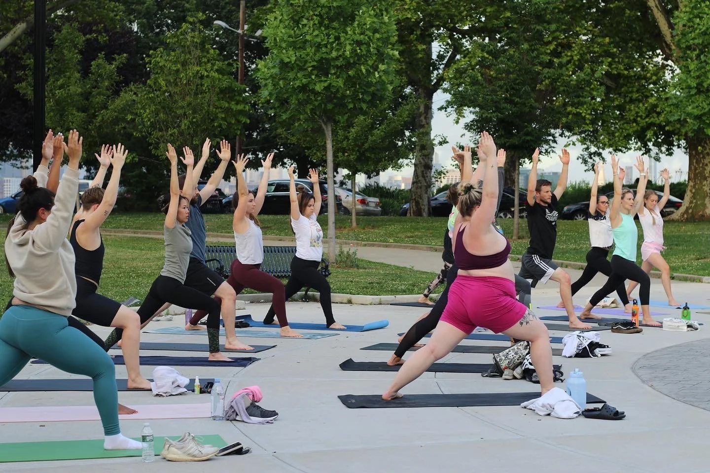 An outdoor yoga class practicing lunges with arms raised among the park’s trees